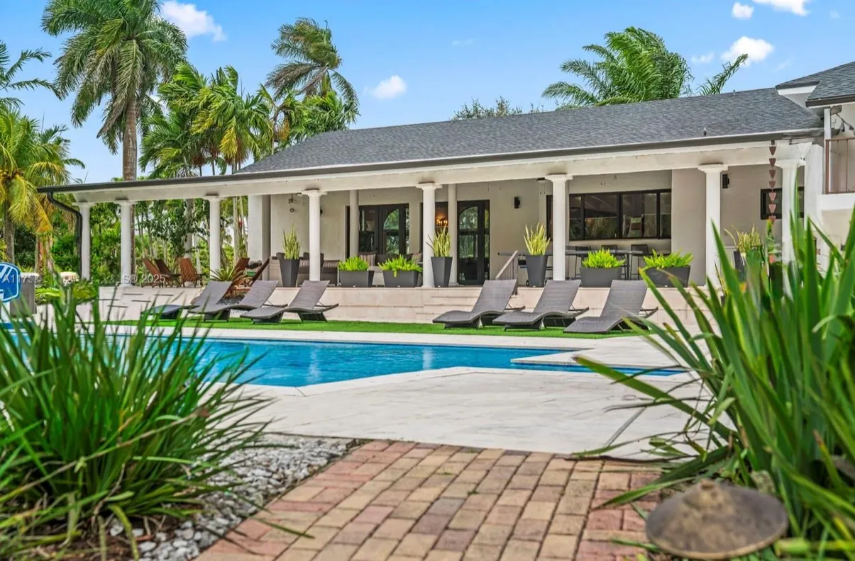 The outdoor area showcases a spacious patio with lounge chairs, a sparkling swimming pool, lush tropical landscaping, and a covered terrace supported by white columns.