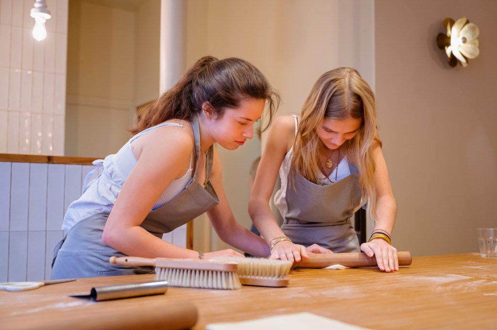 Two people in aprons work together rolling out dough on a wooden table in a kitchen setting.