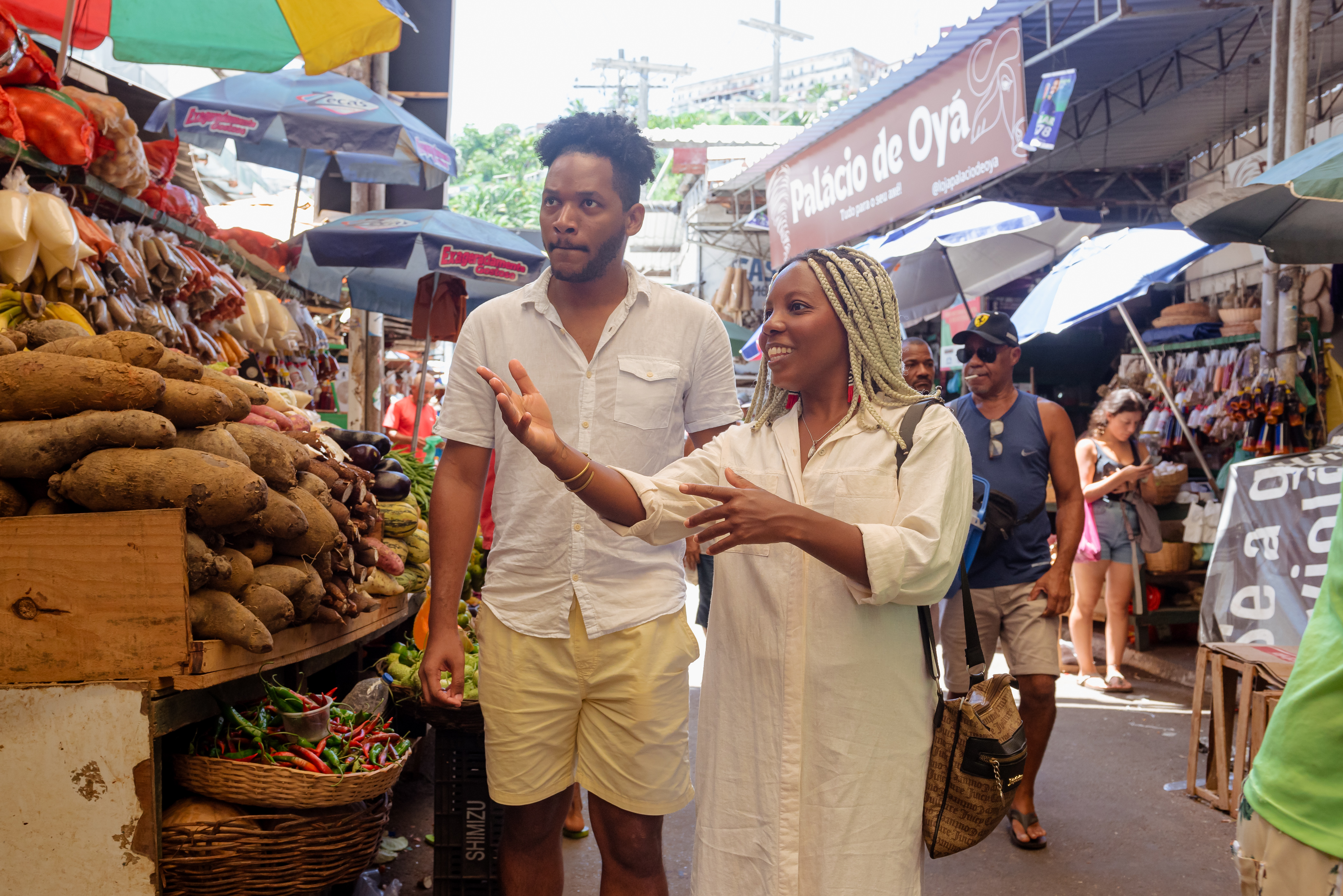 Two people stand and converse at a bustling open-air market, surrounded by piles of fresh produce and vibrant umbrellas.