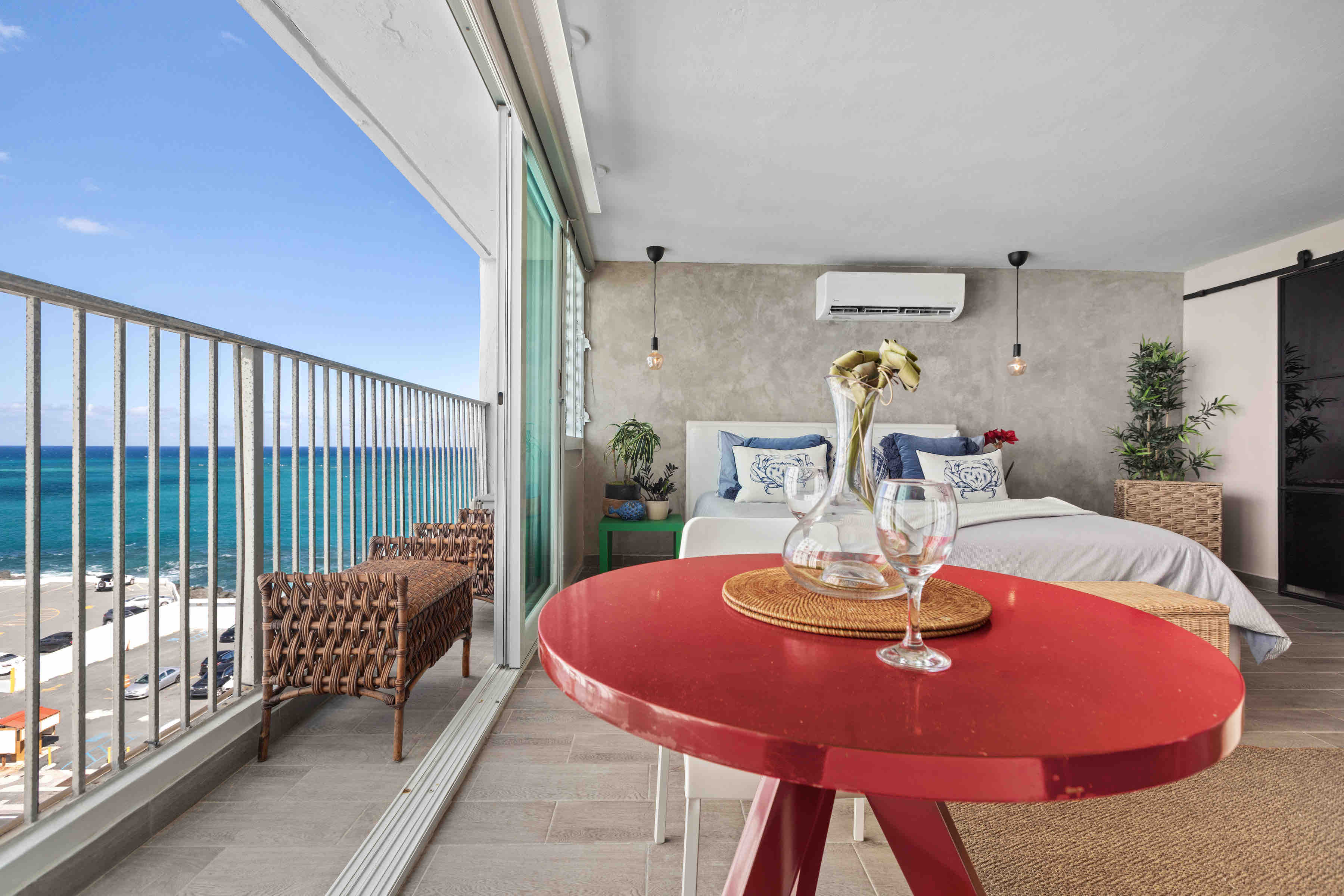 A stylish, modern bedroom opens up to a balcony with wicker seating and panoramic ocean views, featuring a cozy bed and a bright red table set with glassware