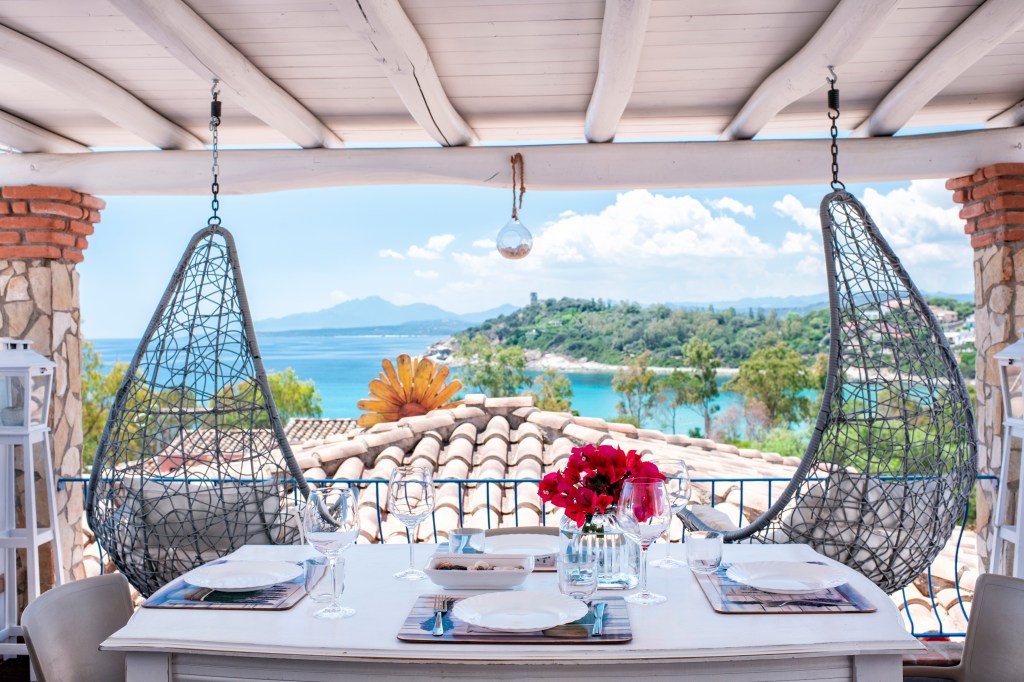 A scenic outdoor dining area with hanging wicker chairs offers panoramic views of turquoise waters, lush greenery, and distant mountains under a sunny sky.