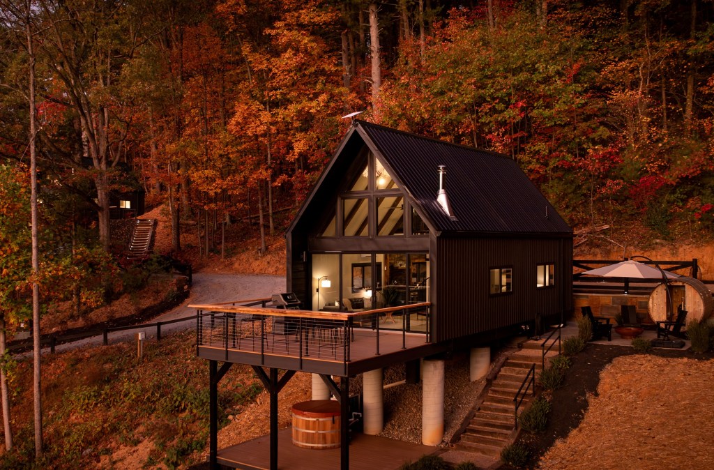 A striking modern cabin with a tall, black roof and outdoor deck nestles into vibrant autumn woods, offering a cozy retreat with a hot tub and sauna.