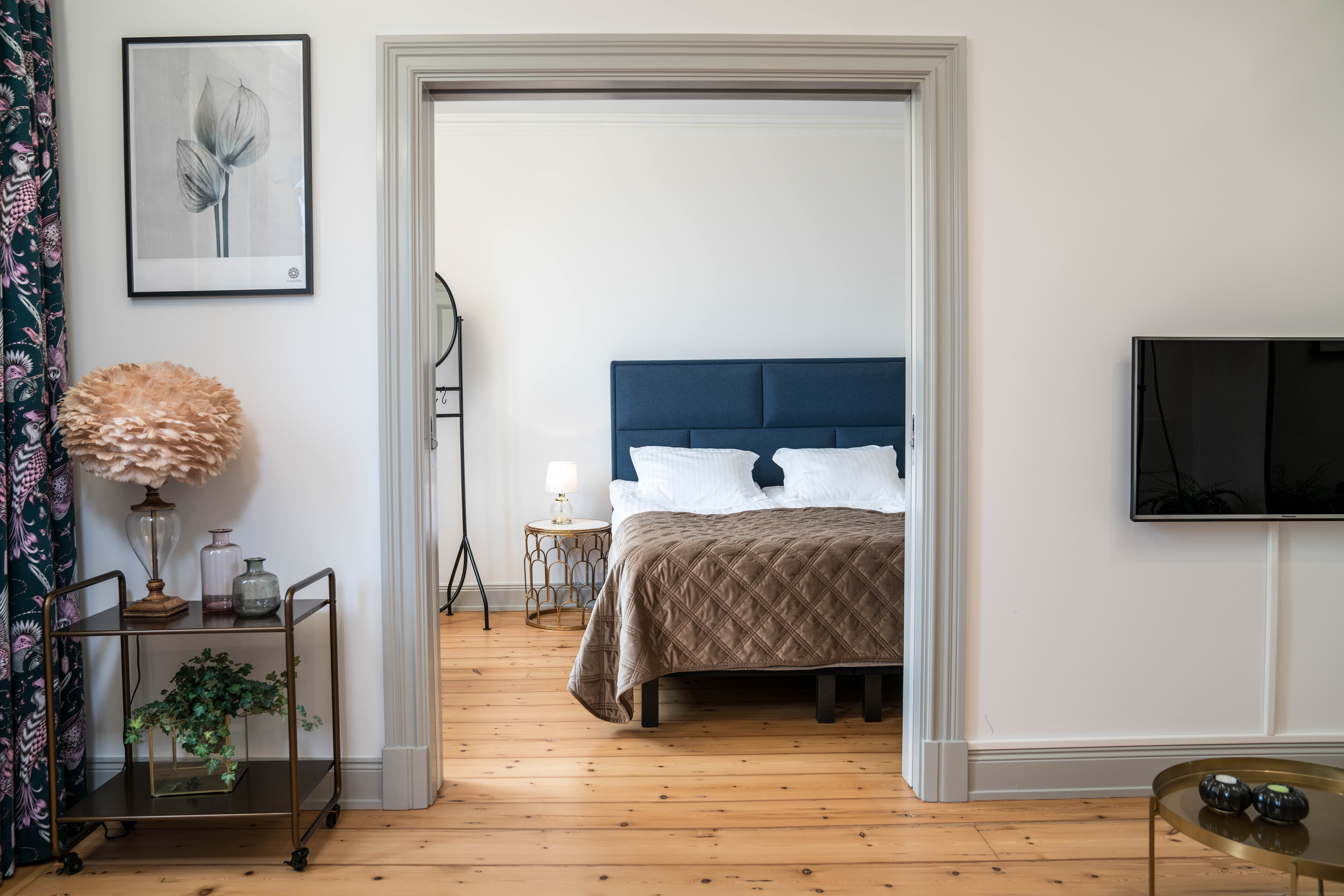 An elegant bedroom with a deep blue headboard and taupe bedding is visible through a wide doorway, complemented by chic decor and soft lighting.