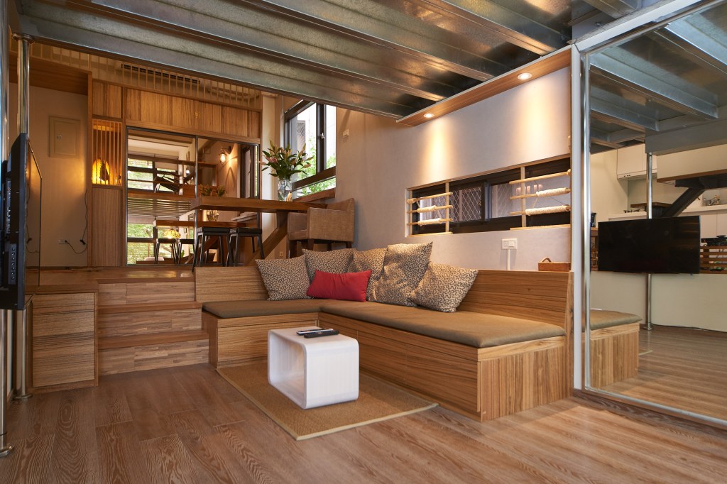 A modern, cozy living room features a wooden sectional couch with patterned cushions, a low white table, and an elevated dining area in the background.