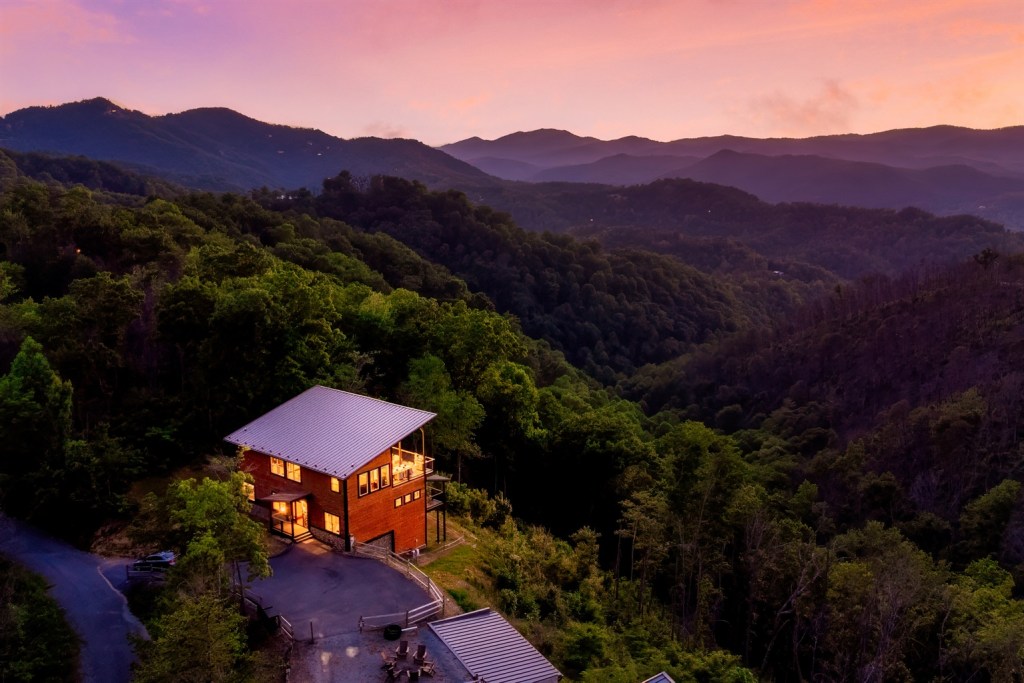 A cozy cabin with glowing windows nestles among lush green mountains, overlooking a breathtaking sunset sky painted in shades of pink and orange.