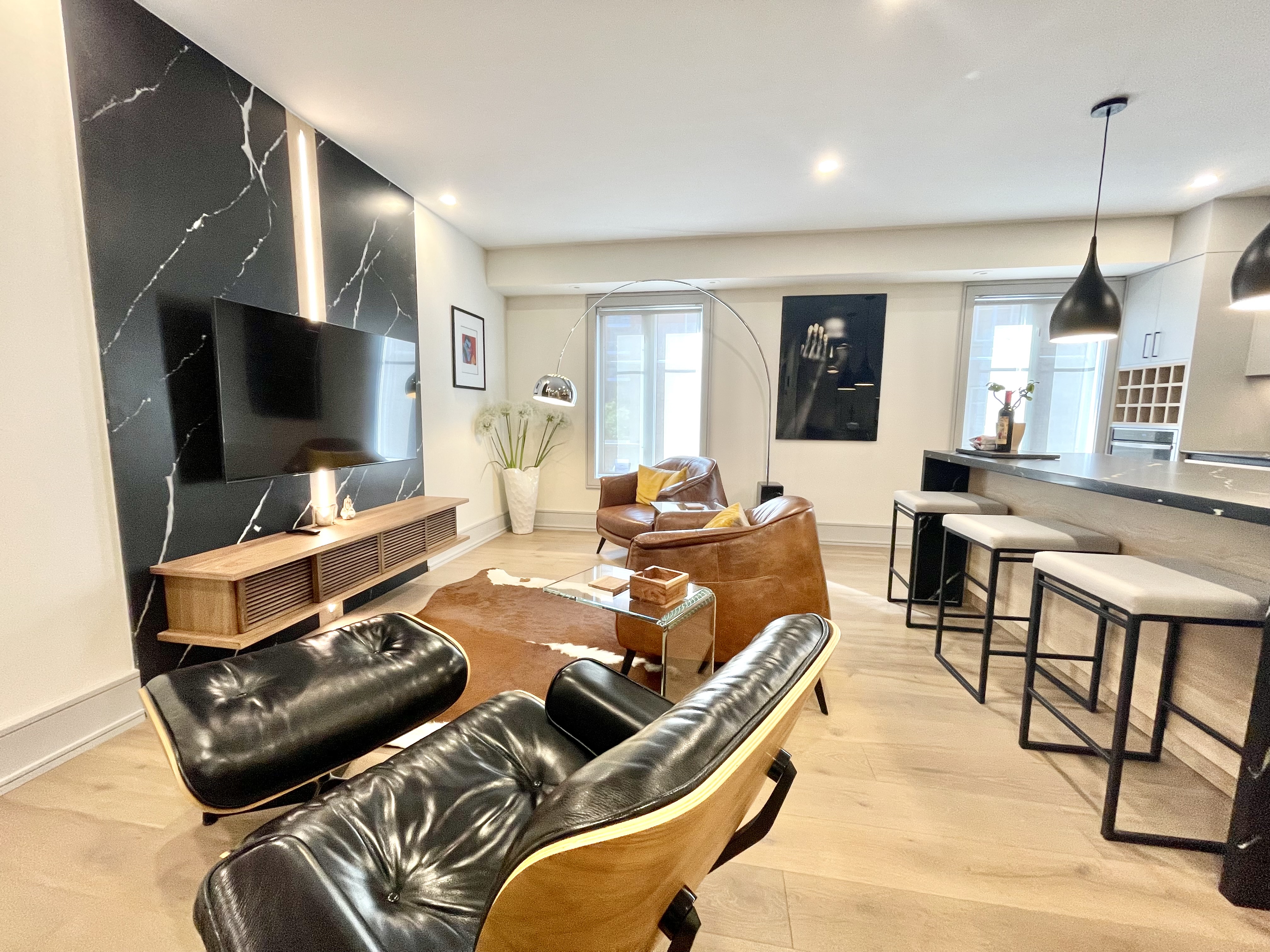 The living room features modern décor with sleek black marble accents, tan leather chairs, a stylish lounge chair, and an open kitchen with bar stools under pendant lighting.