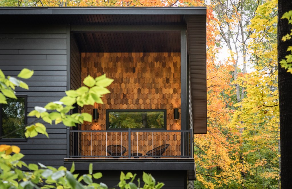 A modern house features a balcony with black railings and honeycomb wood paneling, surrounded by vibrant autumn trees.