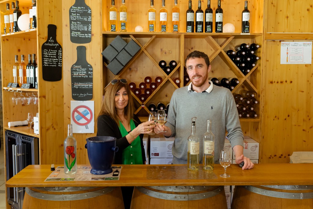 Two people clink glasses behind a wooden bar, surrounded by wine bottles and rustic shelves in a cozy tasting room.