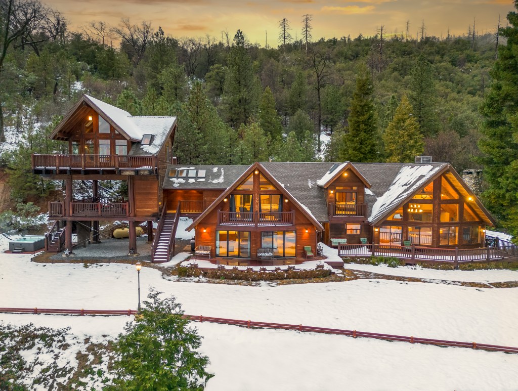 A large, warmly-lit wooden lodge stands amidst snow-covered grounds and pine trees, glowing with inviting light at dusk.